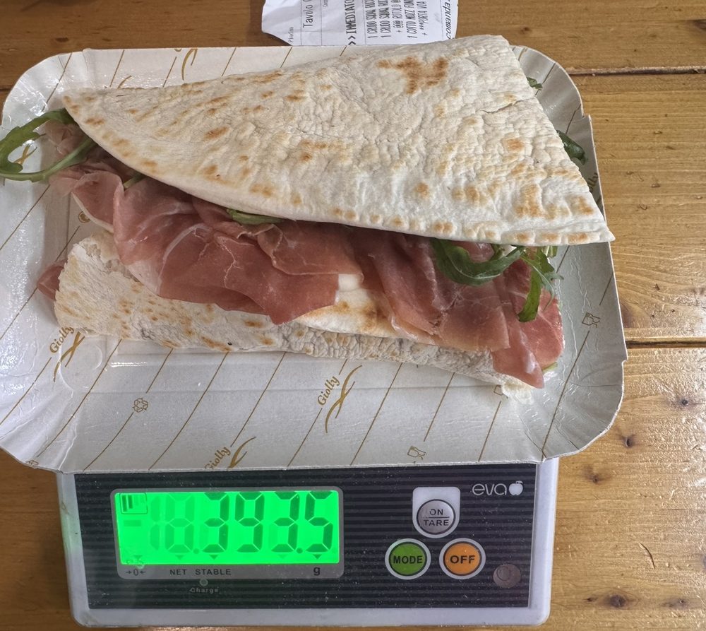 La piada MAXI di Fam da Lóv con crudo e rucola in una vaschetta sulla bilancia: il display segna 393,5 grammi.